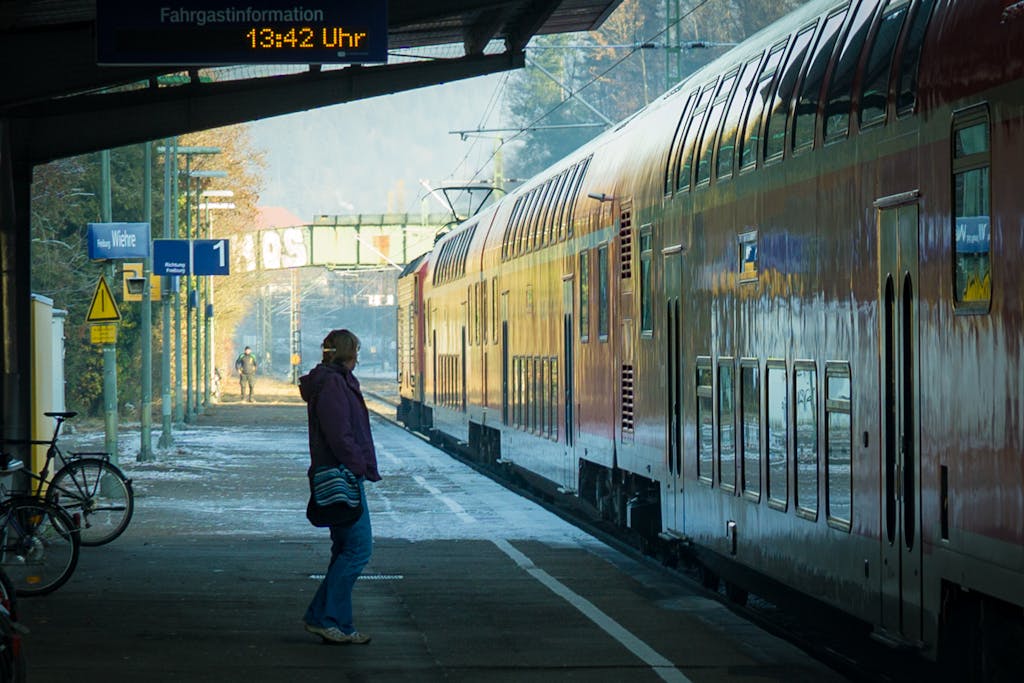 A person waits beside a train at a sunny outdoor station.